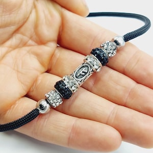 Puede incluir: Un collar de cordón negro con un dije de plata con un símbolo y cuentas de cristal negro y transparente.