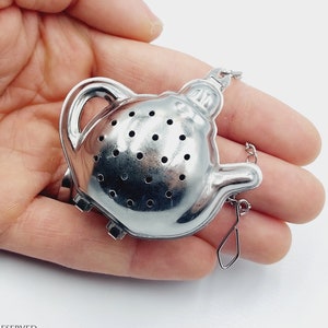 Może przedstawiać: Srebrny zaparzacz do herbaty w kształcie czajniczka z łańcuszkiem i małą zawieszką. Zaparzacz ma uchwyt i dzióbek i jest trzymany w dłoni na białym tle. Zaparzacz jest wykonany z metalu i ma małe otwory.