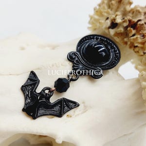 Op de afbeelding: Een zwarte metalen broche met een ovaal frame, een gladde zwarte cabochon, een gefacetteerde zwarte kraal en een vleermuisvormige bedel. De tekst LUCIFEROTHICA staat in het wit gedrukt op de broche die op een textuurrijk bot is geplaatst.
