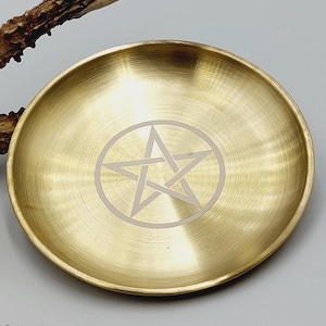 May include: A circular brass dish with a polished metallic finish featuring a centered engraved pentagram symbol. The dish is positioned next to a textured, brown, branch-like object that extends from the upper left corner of the frame.