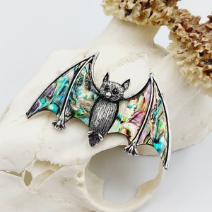 Może przedstawiać: Srebrna broszka w kształcie nietoperza z tęczowymi skrzydłami z muszli abalone. Nietoperz ma szczegółową twarz i jest umieszczony na białym tle.