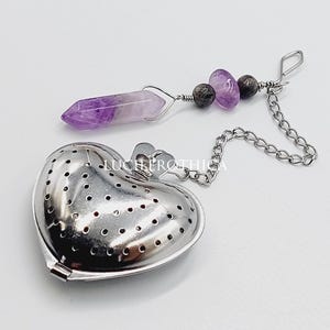 May include: A heart-shaped tea infuser made of silver-colored metal. It has a chain and is adorned with purple and gray gemstones. The infuser is designed for steeping loose leaf tea.