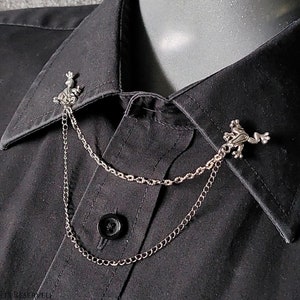 Op de afbeelding: Een zilveren ketting met twee bedels in de vorm van een kikker eraan bevestigd. De ketting ligt over een zwart overhemd met kraag.