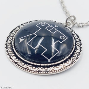 May include: A silver pendant necklace with a black circular charm featuring a white geometric design. The charm has a decorative silver border.