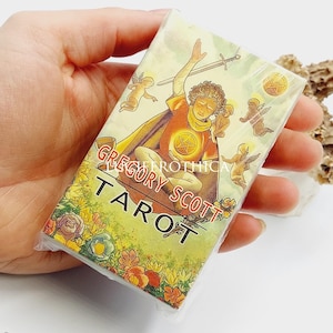 Op de afbeelding: Een pak Gregory Scott Tarotkaarten, vastgehouden in een hand. De kaartkunst toont een figuur met een zwaard, cherubs en bloemmotieven. De tekst "Gregory Scott Tarot" is zichtbaar.
