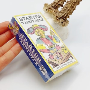 Op de afbeelding: Een "Starter Tarot Deck" doos met een blauw en geel ontwerp. De doos heeft een illustratie van een persoon met een hoed en kleurrijke kleding. De tekst "Starter Tarot Deck" staat op de doos. Het deck is verpakt in transparant plastic.
