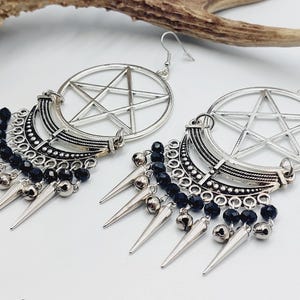 May include: A pair of silver-toned earrings with a pentagram design, crescent moon accents, and dangling spikes. Black beads add detail to the earrings. The earrings are displayed on a white surface.