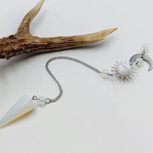 Könnte beinhalten: Ein Pendel an einer silberfarbenen Metallkette mit einer glatten, konischen Spitze aus irisierendem weißem Stein. Die Kette ist mit einem sonnenförmigen Anhänger, einem Mondsichel-Anhänger und kleinen, runden, durchscheinenden weißen Perlen verziert.