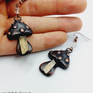 Puede incluir: Un par de pendientes en forma de hongo con un acento de cristal transparente. Los pendientes están hechos de arcilla negra y marrón con detalles en cobre.