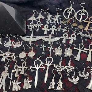 Puede incluir: Una colección de pendientes de metal plateado con motivos egipcios antiguos como ankhs, escarabajos, cobras, deidades aladas y el ojo de Horus, junto a una pirámide negra y el texto Luciferthica.com All Rights Reserved.