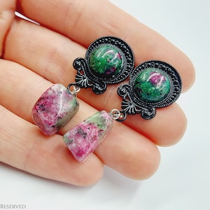 Op de afbeelding: Een paar oorhangers met groene en roze edelsteen druppels. De oorbellen hebben zwarte metalen instellingen met ingewikkelde ontwerpen.