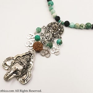 Op de afbeelding: Een zilveren ketting met een groene en zwarte kralenketting. De ketting heeft een zilveren hanger met een olifantenkop en een zilveren bedel met het Om-symbool. De hanger is versierd met groene stenen en een bruine kraal.