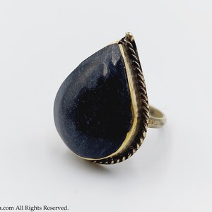 Pode incluir: Um anel em forma de lágrima com uma pedra preta engastada em uma banda de metal dourado. A banda tem um design de corda.