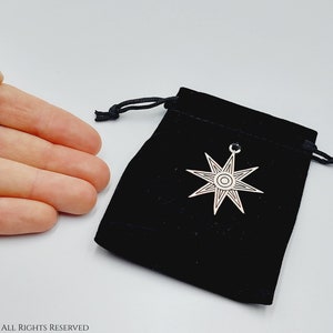 Pode incluir: Um pingente de estrela de oito pontas de prata com um círculo no centro, descansando em uma bolsa de veludo preto com cordão.