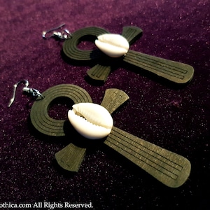 May include: A pair of wooden Ankh earrings with a white cowrie shell accent. The earrings are dark brown and have a textured finish.
