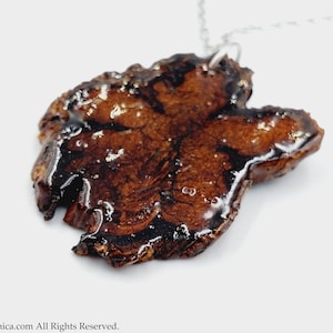 Può includere: Un ciondolo in resina marrone e nero con una forma organica unica, appeso a una catena d'argento. Il ciondolo ha una finitura lucida e sembra essere fatto di un materiale naturale.