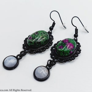 Puede incluir: Un par de pendientes colgantes con cabujones de piedras preciosas verdes y rosas engastados en marcos de metal negro. Los pendientes tienen una piedra preciosa redonda azul más pequeña que cuelga debajo de la piedra preciosa más grande.