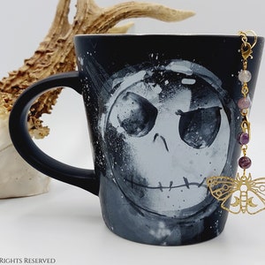 Könnte beinhalten: Schwarze Keramiktasse mit einem weißen Jack Skellington-Design. Eine goldene Kette mit lila und rosa Perlen und einem goldenen Schmetterlingsanhänger hängt an der Tasse.