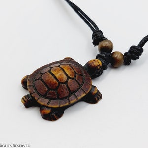 Può includere: Una collana con un ciondolo a forma di tartaruga marrone su un cordoncino nero e perline di legno. La tartaruga è scolpita con dettagli sul guscio.