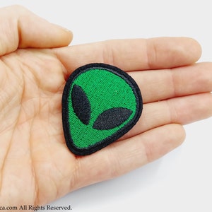 Può includere: Toppa ricamata con il viso di un alieno verde con contorno nero. La toppa è su una mano.