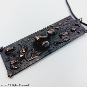 May include: A rectangular pendant necklace with a textured, dark brown surface and a black cord. The pendant has raised, irregular shapes and a bronze-colored accent. The overall design is artistic and unique.