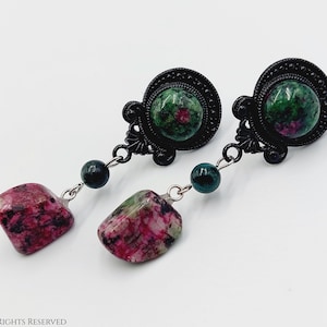 Op de afbeelding: Een paar oorhangers met zwarte metalen instellingen en groene en roze edelsteenaccenten.