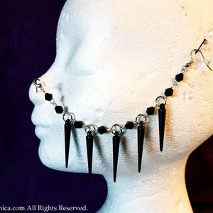 Op de afbeelding: Een zilveren neusring met een ketting, zwarte kralen en zwarte spike-bedels. De ketting is bevestigd aan een zilveren oorhaak.