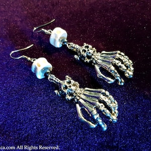 Op de afbeelding: Een paar zilveren skelethand-oorbellen met witte kralenaccenten. De oorbellen hangen en hebben een schedel bovenaan de hand.