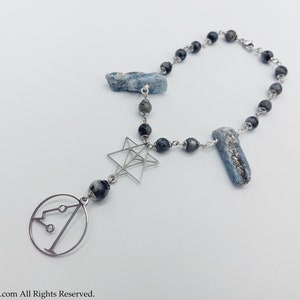 Pode incluir: Um colar de prata com contas pretas e detalhes de cristal azul. O colar apresenta um pingente de estrela de prata e um pingente de círculo de prata com um design geométrico.