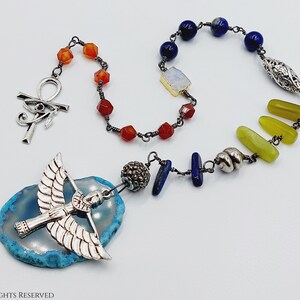 Op de afbeelding: Een handgemaakte ketting met een zilveren Ankh-hanger en een gevleugelde godheid-bedel op een blauwe agaat schijf. De ketting bevat oranje, blauwe, groene en zilveren edelsteen kralen, verbonden door een donkere metalen ketting.