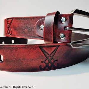 May include: A red leather belt with a silver buckle and a black symbol embossed on the leather. The symbol is a stylized inverted pentagram.