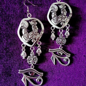 May include: Silver dangle earrings with a detailed design of two figures with headdresses and a dangling eye of Horus charm.