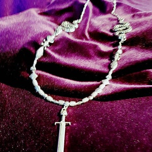 May include: A silver necklace with a sword pendant on a purple background.