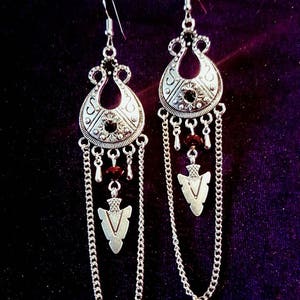 Op de afbeelding: Zilveren oorhangers met een traanvormige hanger, een ketting en een kleine pijlbedel. De oorhangers hebben een accent van donkerrode kralen.