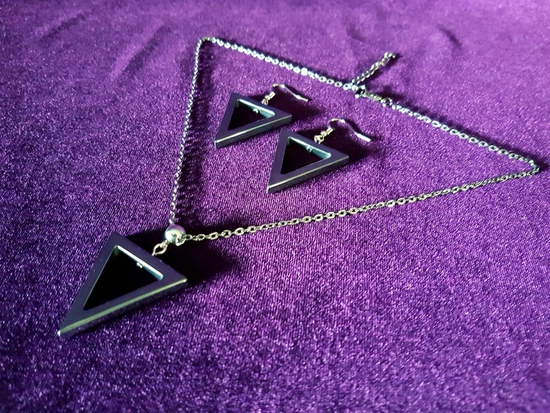 May include: A silver necklace and earrings set with a geometric triangle pendant and matching earrings. The pendant and earrings are hollow and have a black interior.