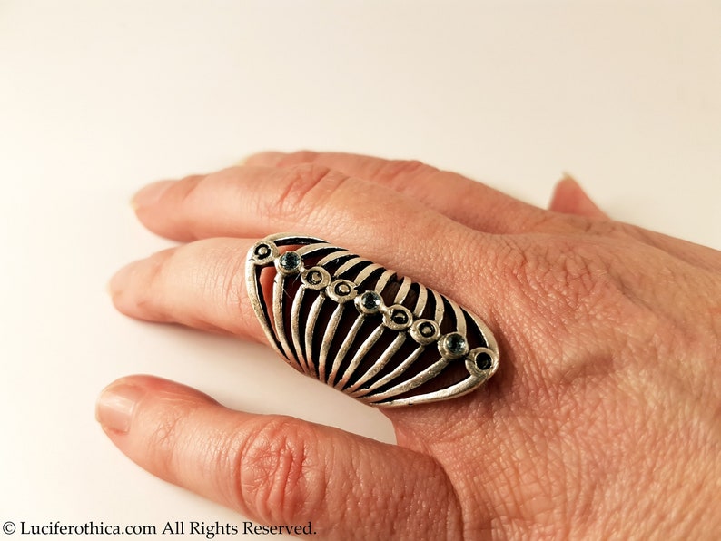Pu&ograve; includere: Un anello d'argento con un design a forma di gabbia e pietre preziose blu. L'anello &egrave; indossato su un dito.