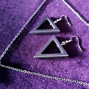 May include: A silver chain necklace with a silver triangle pendant and a pair of silver triangle earrings. The jewelry is displayed on a purple velvet surface.