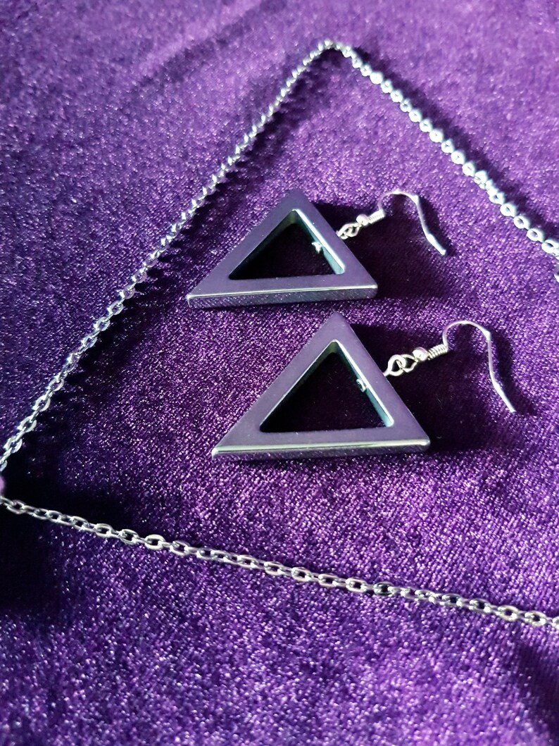 May include: A silver chain necklace with a silver triangle pendant and a pair of silver triangle earrings. The jewelry is displayed on a purple velvet surface.