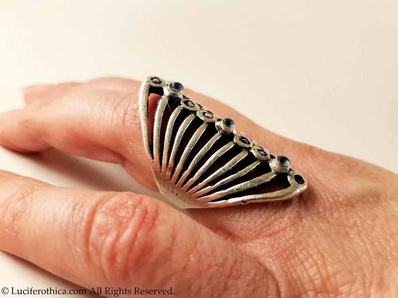 Pu&ograve; includere: Un anello d'argento con un design a forma di ventaglio e pietre preziose blu. L'anello &egrave; indossato su un dito.