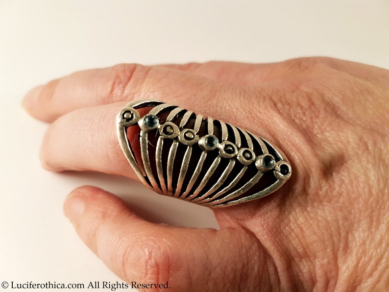 Pu&ograve; includere: Un anello d'argento con un design a gabbia e pietre preziose blu. L'anello &egrave; indossato su un dito.
