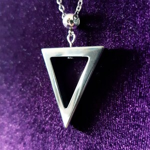 May include: A silver necklace with a triangle pendant. The pendant is hollow and has a geometric design. The necklace is on a purple velvet background.