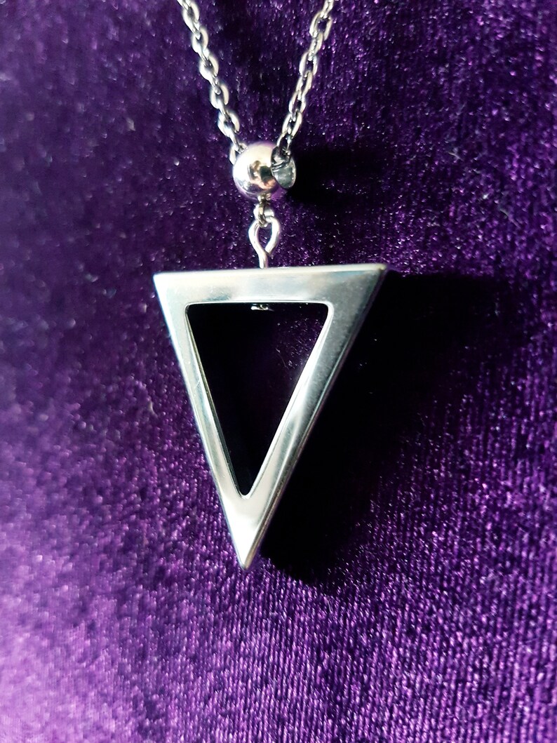 May include: A silver necklace with a triangle pendant. The pendant is hollow and has a geometric design. The necklace is on a purple velvet background.