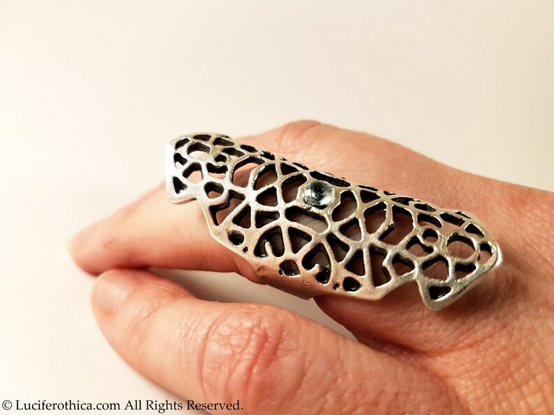 May include: A silver ring with a delicate, intricate design and a small, clear gemstone. The ring is worn on a finger and is a statement piece.