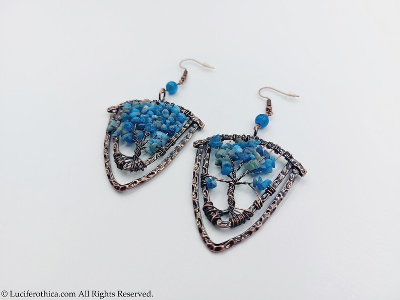 May include: A pair of copper wire earrings with a tree of life design. The tree is made of blue and green gemstone chips. The earrings are in the shape of a triangle.