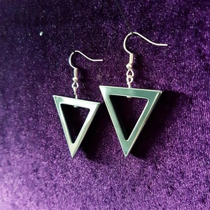 May include: A pair of silver triangle earrings with a black triangle cutout in the center. The earrings are hanging on a purple velvet background.