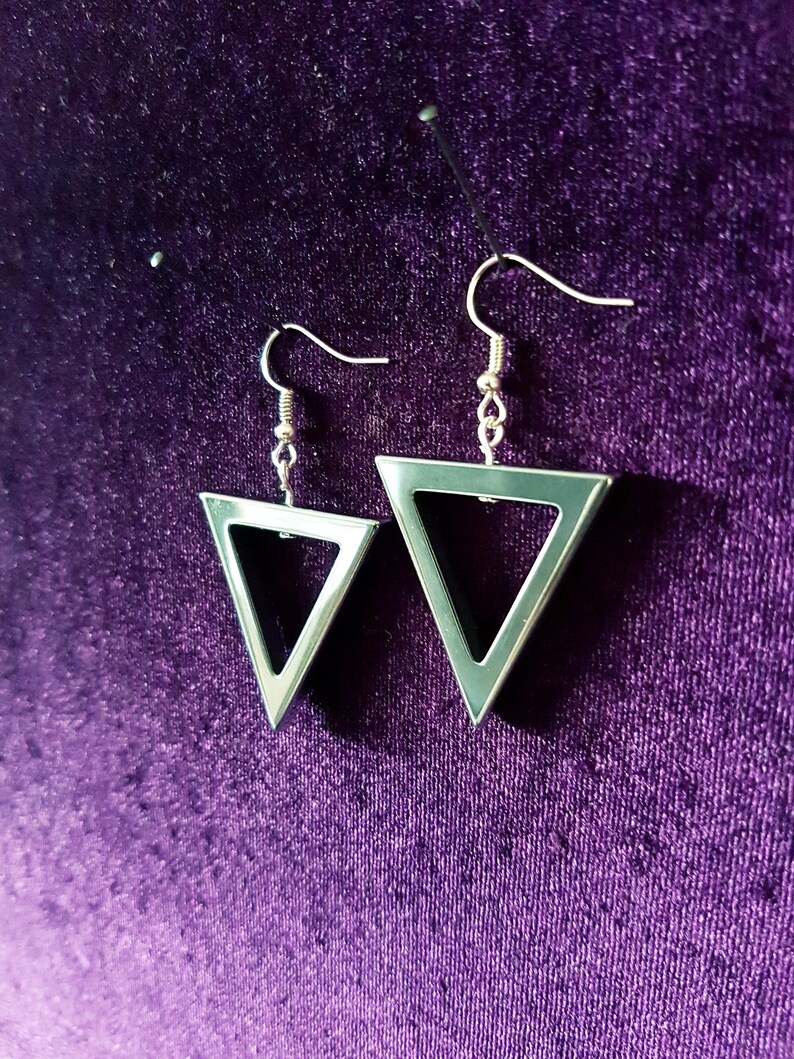 May include: A pair of silver triangle earrings with a black triangle cutout in the center. The earrings are hanging on a purple velvet background.