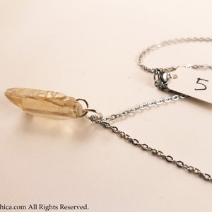 May include: A silver chain necklace with a light brown crystal pendant. The pendant is a long, thin, pointed crystal. The chain is a delicate, cable chain. The necklace is on a white background. The number 5 is written on a white tag.