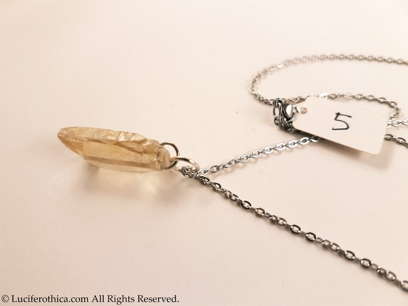 May include: A silver chain necklace with a light brown crystal pendant. The pendant is a long, thin, pointed crystal. The chain is a delicate, cable chain. The necklace is on a white background. The number 5 is written on a white tag.