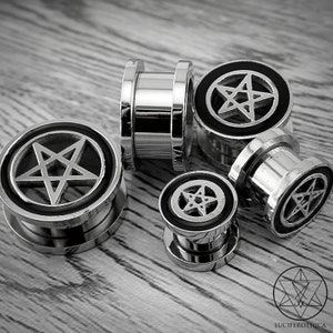 May include: Set of five silver metal ear gauges with black pentagram designs. The gauges are different sizes and are arranged on a wooden surface.