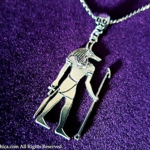 May include: Silver pendant necklace featuring an Egyptian god Anubis with a jackal head, holding a staff. The pendant is on a silver chain and is displayed on a purple velvet background.